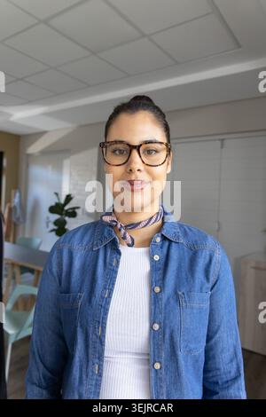 Frau, die im Essbereich zu Hause steht und mit der Kamera zugewandt ist, trägt Denim-Hemd und Schildpatt-Brille. Porträt, Innenraum, Contempo Stockfoto