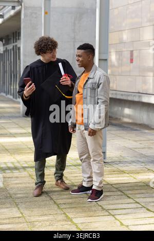 Verschiedene männliche Freunde plaudern auf dem Campus, Abschluss im Kleid mit Diplom, Mütze, Telefon, Brille. Universität, Zeremonie, Feier Stockfoto