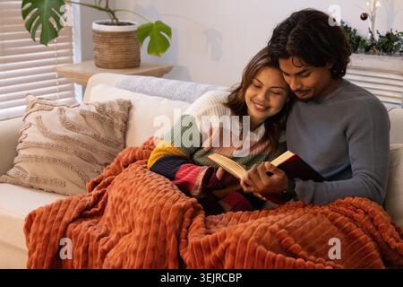 Diverse Paare, die offenes Buch auf einem leichten Sofa lesen, teilen sich Rost-Throw-Frau in gestreiftem Pullover. Gemütlich, Wärme, Decke, Kissen, MON Stockfoto