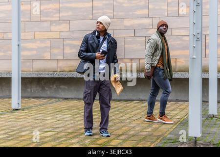 Verschiedene männliche Freunde stehen und gehen auf dem städtischen plaza mit Smartphone und Essensbeutel. Stadtbild, Innenhof, Fußgängerzone Stockfoto