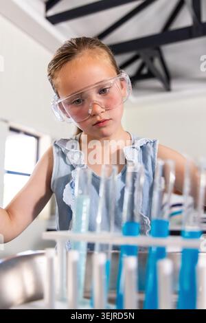 Schulmädchen mit Brille und ärmellosem Oberteil, untersucht Reagenzgläser mit blauer Flüssigkeit im Schullabor. Kind, Schutzbrille, Klasse Stockfoto