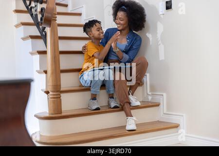 Afroamerikanische Mutter und hispanischer Sohn sitzen zu Hause auf hölzernen Treppen und teilen sich eine blaue Tafel. Familie, Lebensstil, offen, Stockfoto