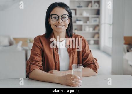 Lächelnde junge Frau, die zu Hause ein Glas Wasser hält und am Tisch im hellen Wohnzimmer sitzt Stockfoto