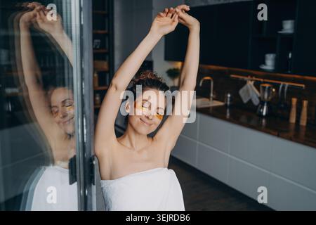 Entspannte junge Frau, die die Arme am Morgen mit Augenaufnäher und Handtuch zu Hause dehnt Stockfoto