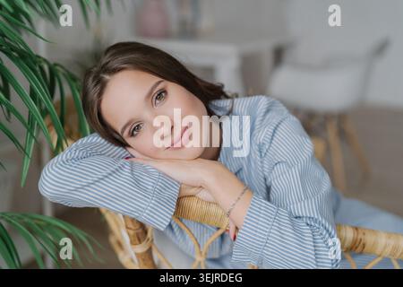 Entspannte junge Frau ruht Kopf auf Weidenstuhl zu Hause in der Nähe von grüner Pflanze, die blaues gestreiftes Hemd in ruhiger natürlicher Inneneinrichtung trägt Stockfoto