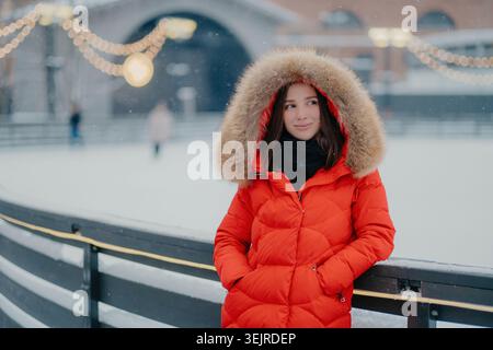 Nachdenkliche junge Frau in rotem Wintermantel, die bei Schneefall neben der Eislaufbahn steht und festliche Stadtlichter hinter sich hat Stockfoto