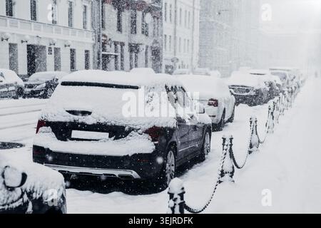 Schneebedeckte Autos parkten auf der City Street bei starkem Winterschnee mit städtischen Gebäuden, Gehsteigen und Copy Space Stockfoto