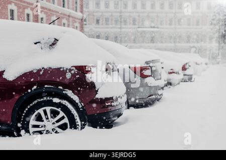 Schneebedeckte Autos auf dem Parkplatz während des schweren Wintersturms mit fallendem Schnee, städtischen Gebäuden und kaltem Wetter Reisehintergrund Stockfoto