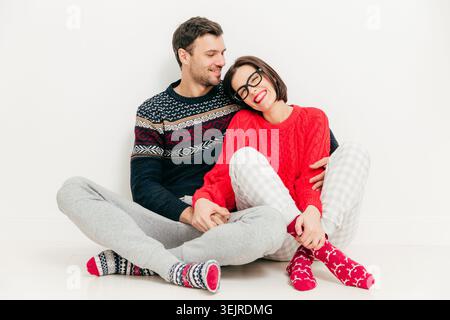 Glückliches junges Paar, das zu Hause auf dem Boden sitzt, in warmen Pullis, die zusammen vor sauberem Hintergrund der weißen Wand lächeln Stockfoto