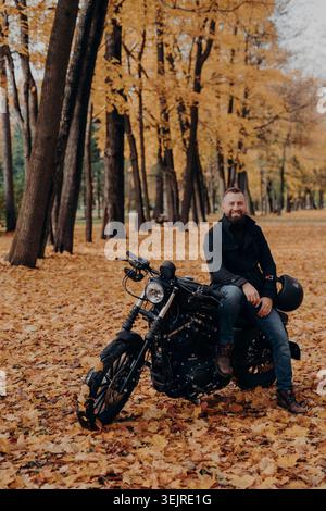 Fröhlicher Mann mittleren Alters, der auf schwarzem Motorrad im Autumn Park sitzt, umgeben von gefallenen Blättern während einer Fahrt im Freien Stockfoto