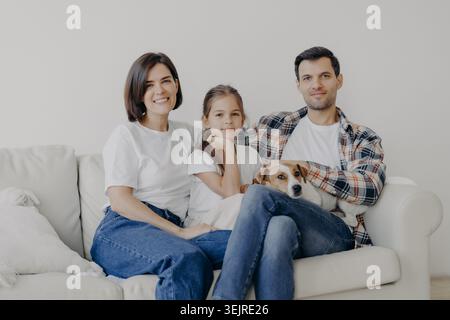 Lächelnde Millennial-Familie mit einer jungen Tochter und einem kleinen Hund, die zusammen auf dem weißen Sofa in einem hellen Wohnzimmer sitzen Stockfoto