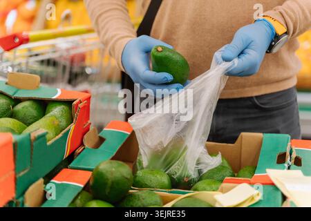 Nahaufnahme von Handschuhen, die frische Avocados in der Sektion für Supermärkte wählen, während grüne Früchte in Plastiktüte gelegt werden Stockfoto