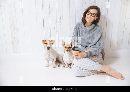 Fröhliche Millennial-Frau sitzt auf dem Boden mit zwei kleinen Hunden und Kaffeetasse zu Hause vor der weißen Holzwand Stockfoto