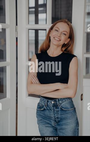 Selbstbewusste rothaarige Frau, die zu Hause mit überkreuzten Armen steht und in lässigem Schwarz-Top und blauer Jeans natürlich lächelt Stockfoto