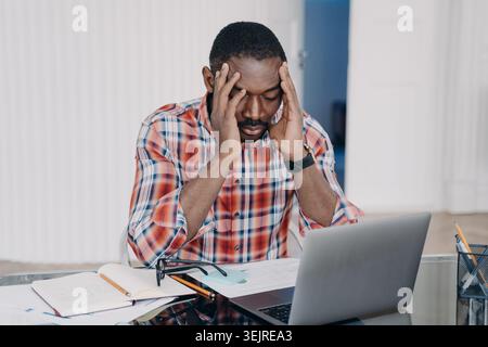 Gestresster Schwarzer Mann, der im Home Office an einem Laptop arbeitet und den Kopf in den Händen hält, während er sich von Papieren und Notizen überwältigt fühlt Stockfoto