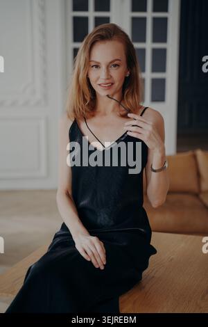 Elegante rothaarige Frau, die auf Holztisch sitzt, Brillen hält und in stilvollem schwarzen Kleid im Innenraum lächelt Stockfoto