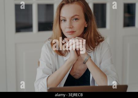 Nachdenkliche Millennial-Frau, die im hellen Home Office an einem Laptop arbeitet und am Schreibtisch sitzt, mit Händen unter dem Kinn und beruhigendem FAC sitzt Stockfoto