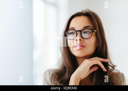 Nachdenkliche junge Frau, die eine Brille in der Nähe eines hellen Fensters trägt, mit Hand unter dem Kinn in weichem natürlichem Licht und ruhiger Indoor-Stimmung Stockfoto