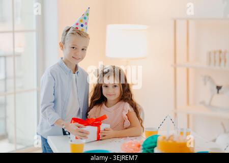 Glückliche Kinder feiern zu Hause Geburtstagsfeier mit Geschenkbox in der Nähe von Kuchentassen und farbenfrohen Dekorationen im hellen Raum Stockfoto