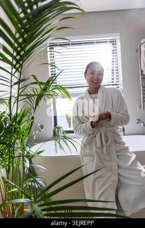 Asiatische Frau im Alter von 20 Jahren, die in einem hellen Badezimmer mit Pflanzen auf dem Wannenrand sitzt und weißen Bademantel trägt. Bademantel, freistehend, Spa, grün, Stockfoto