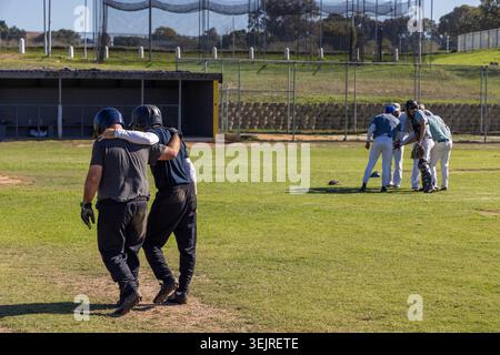 Männliche Mannschaftskameraden, darunter ältere Spieler, die Arm in Arm auf dem lokalen Baseballfeld mit Helmen unterwegs sind. Kameradschaft, Teamwork, draußen, sonnig, Dugout, fen Stockfoto