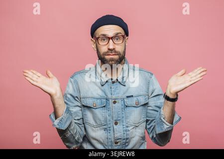 Verwirrter bärtiger Mann zuckt mit erhobenen Händen an der pinkfarbenen Studiowand Stockfoto