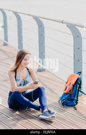 Junge Frau, die nach dem Training im Freien mit Smartphone und Ohrhörern auf der Promenade sitzt Stockfoto