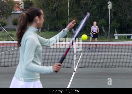 Erwachsene Spielerinnen in Sportbekleidung, die auf dem Platz Tennis schwingen und beim gelben Ball über dem Netz spielen. Outdoor, dynamisch, unscharf, ATH Stockfoto