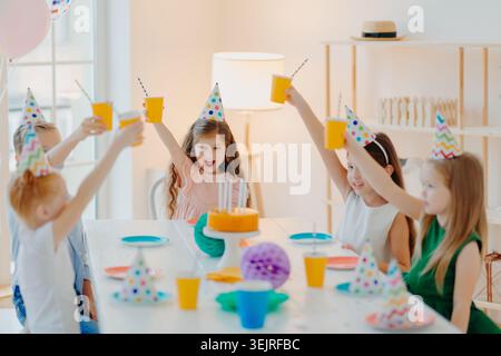Kinder in Partyhüten heben während der Hausfeier Becher um Geburtstagskuchen Stockfoto