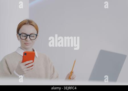 Junge Frau mit Brille liest das Smartphone, während sie Notizen neben dem Laptop im Home Office macht Stockfoto