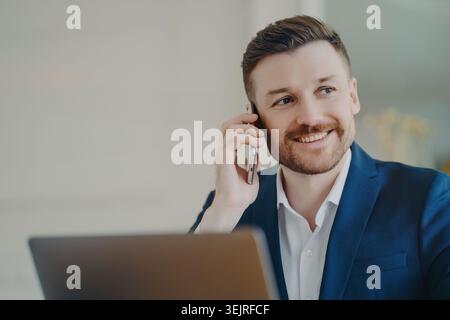 Ein lächelnder junger Geschäftsmann spricht über ein Smartphone, während er im modernen Büro mit einem Laptop arbeitet Stockfoto