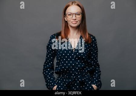 Junge rothaarige Frau in Brille steht selbstbewusst vor grauer Studiowand Stockfoto
