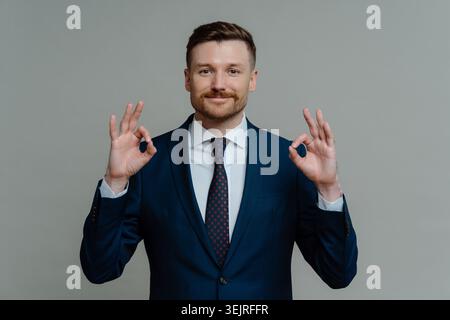 Geschäftsmann im blauen Anzug zeigt eine gute Geste mit beiden Händen gegen die graue Wand Stockfoto