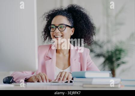 Lächelnde Frau in rosa Blazer arbeitet am Computer in einem hellen, modernen Büro Stockfoto