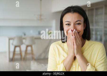 Junge Frau, die den Mund bedeckt, mit beiden Händen, die vor Überraschung lachen, im hellen modernen Innenraum mit gelbem Oberteil Stockfoto