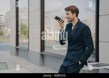 Der junge Geschäftsmann spricht vor dem modernen Bürogebäude in das Smartphone, während der Laptop auf dem Tisch liegt Stockfoto