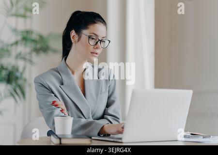 Junge Geschäftsfrau in grauem Blazer und schwarzer Brille, die am Schreibtisch mit Papierkaffeetasse im hellen, modernen Büro auf einem Laptop schreibt Stockfoto