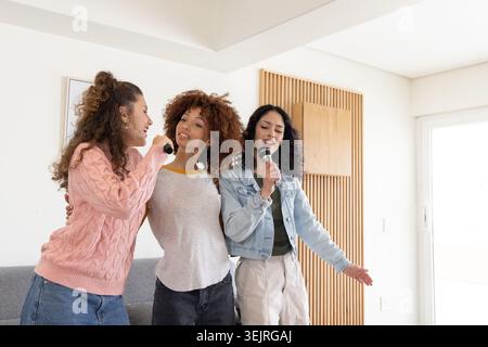 Drei Freundinnen stehen nebeneinander und singen in Handmikrofonen in der Nähe des grauen Sofas zu Hause. Frauen, Trio, Musik, spielen Stockfoto