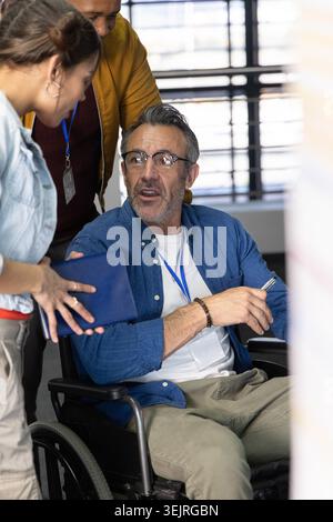 Kollegen treffen sich im Büro mit einem Mann im Rollstuhl, der Stift, Tablet, Schlüsselband hält. Zusammenarbeit, Brainstorming, Konf Stockfoto