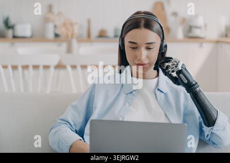 Junge Frau mit Armprothese verwendet Laptop und Headset, während sie von zu Hause aus arbeitet Stockfoto