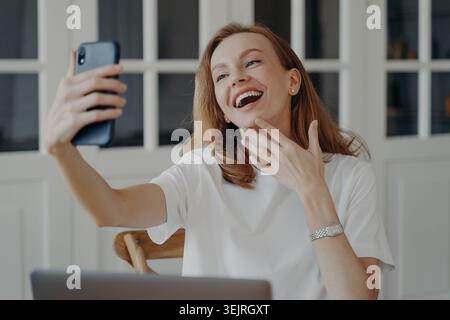 Junge rothaarige Frau lächelt, während sie im Heimbüro neben dem Laptop ein Smartphone-Selfie macht Stockfoto