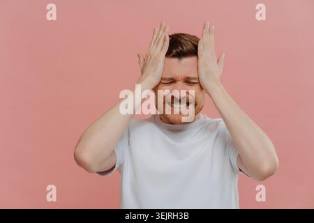 Gestresster Mann, der seinen Kopf mit beiden Händen gegen die rosa Studiowand hält Stockfoto