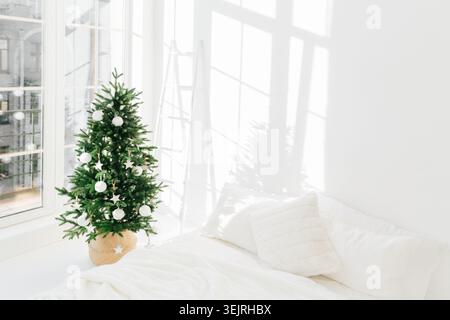 Der geschmückte Weihnachtsbaum steht neben dem weißen Sofa in einem hellen, minimalistischen Wohnzimmer Stockfoto