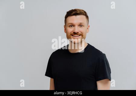 Ein lächelnder junger Mann in schwarzem T-Shirt steht an der normalen Studiowand Stockfoto