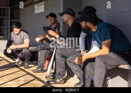 Eine Gruppe von fünf männlichen Baseballspielern sitzt und hockt sich in der Dugout, hält Fledermaushandschuh Baseball. Team, Bank, Feld, Sonnenlicht, SHA Stockfoto