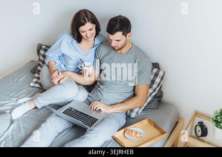 Ein junges Paar benutzt einen Laptop zusammen im Bett mit Kaffee und Frühstückstablett zu Hause Stockfoto