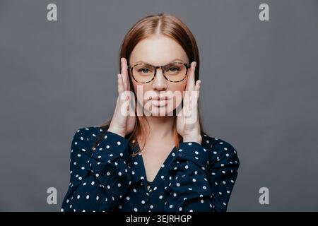 Die rothaarige Frau passt die Brille an, während sie die Kamera im grauen Studio-Porträt betrachtet Stockfoto