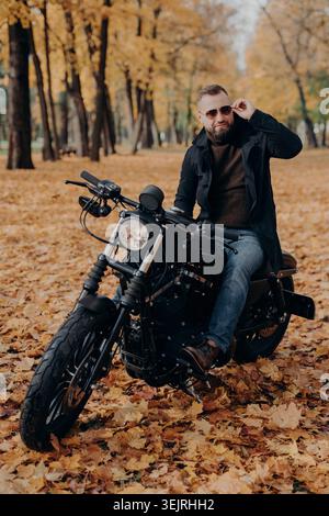 Bärtiger Mann mit Sonnenbrille sitzt auf schwarzem Motorrad im Herbstpark Stockfoto