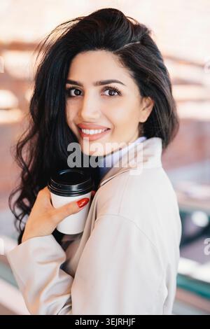 Die junge Frau lächelt, während sie eine Tasse Kaffee zum Mitnehmen auf dem städtischen Balkon hält Stockfoto