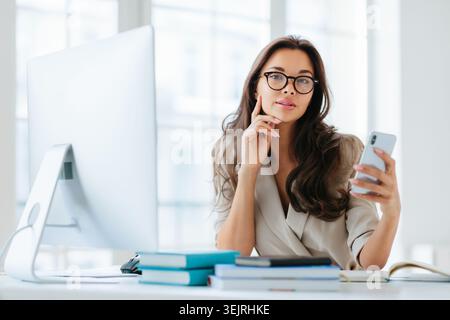 Die junge Geschäftsfrau mit Brille hält das Smartphone am Schreibtisch in einem hellen Büro Stockfoto